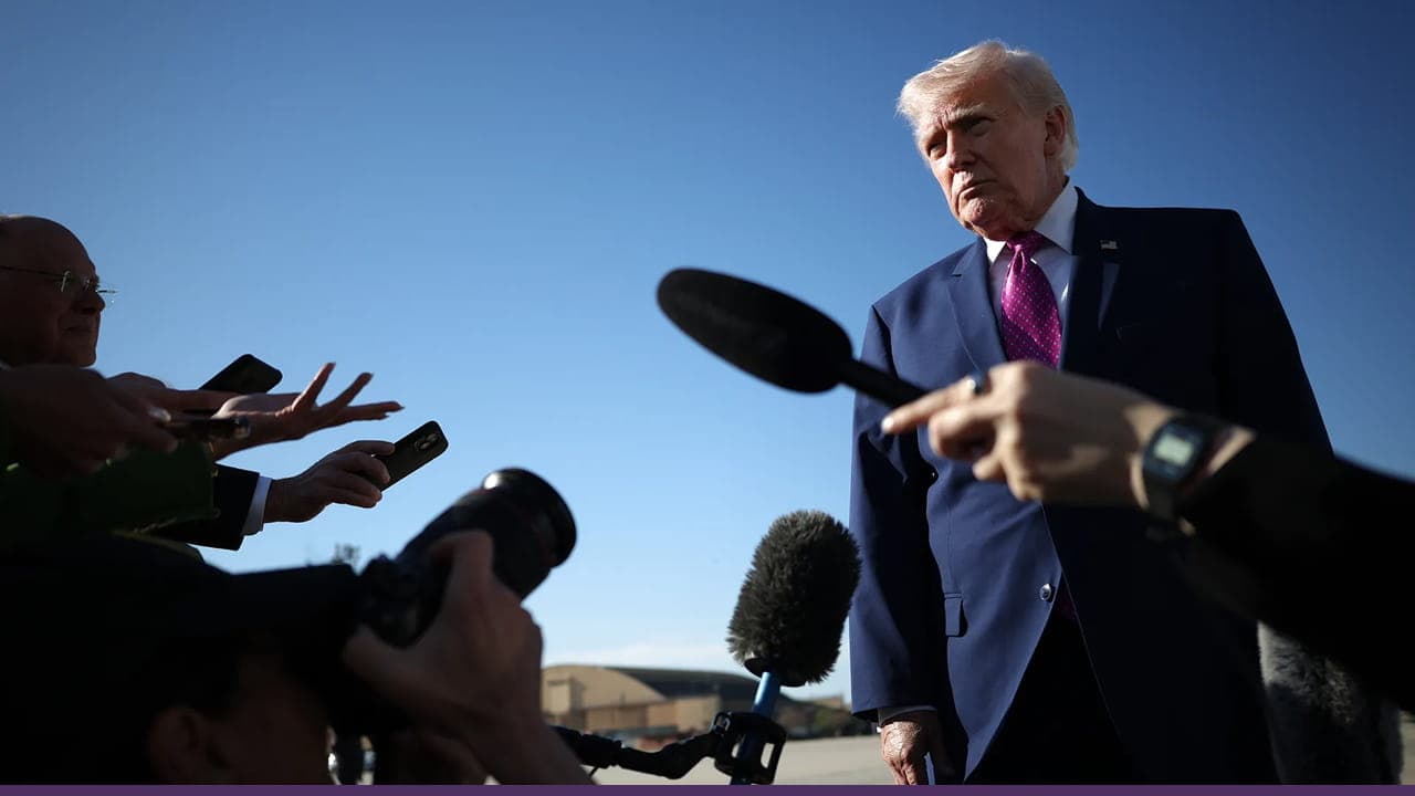 A man in a suit stands outdoors under a clear blue sky while speaking to reporters, with several microphones, cameras, and hands holding recording devices extended toward him during a press interaction.