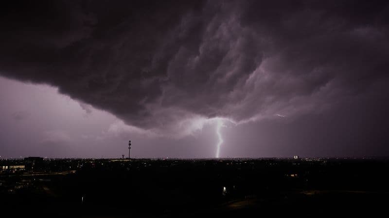 Dark, heavy storm clouds loom over a city at night as a bright lightning bolt strikes down to the ground in the distance, illuminating the skyline below.
