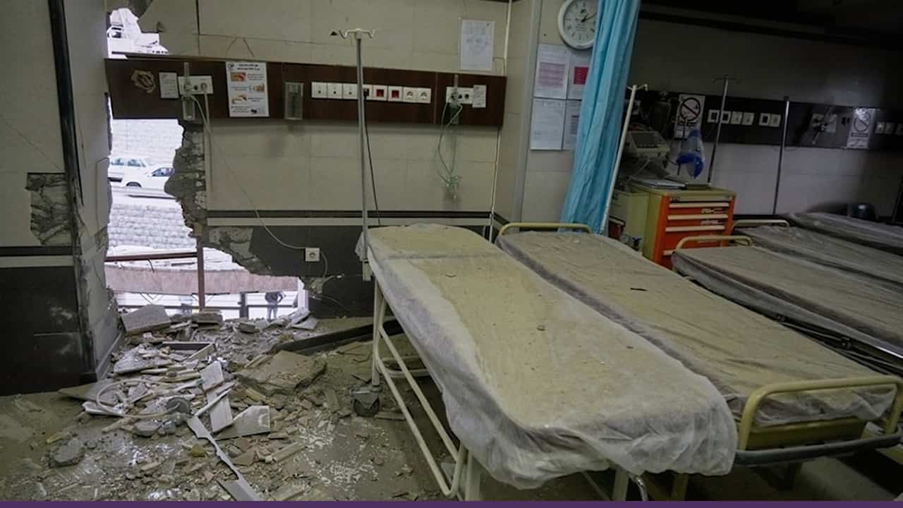 A damaged hospital room with two empty beds covered in dust, debris scattered across the floor, and a large hole in the wall exposing the outside.