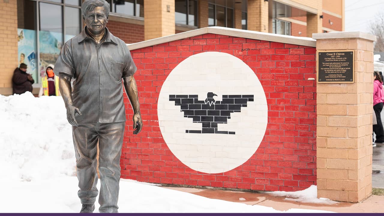A statue of Cesar Chavez stands beside a mural featuring the United Farm Workers eagle symbol, set against a brick wall in a public outdoor space.