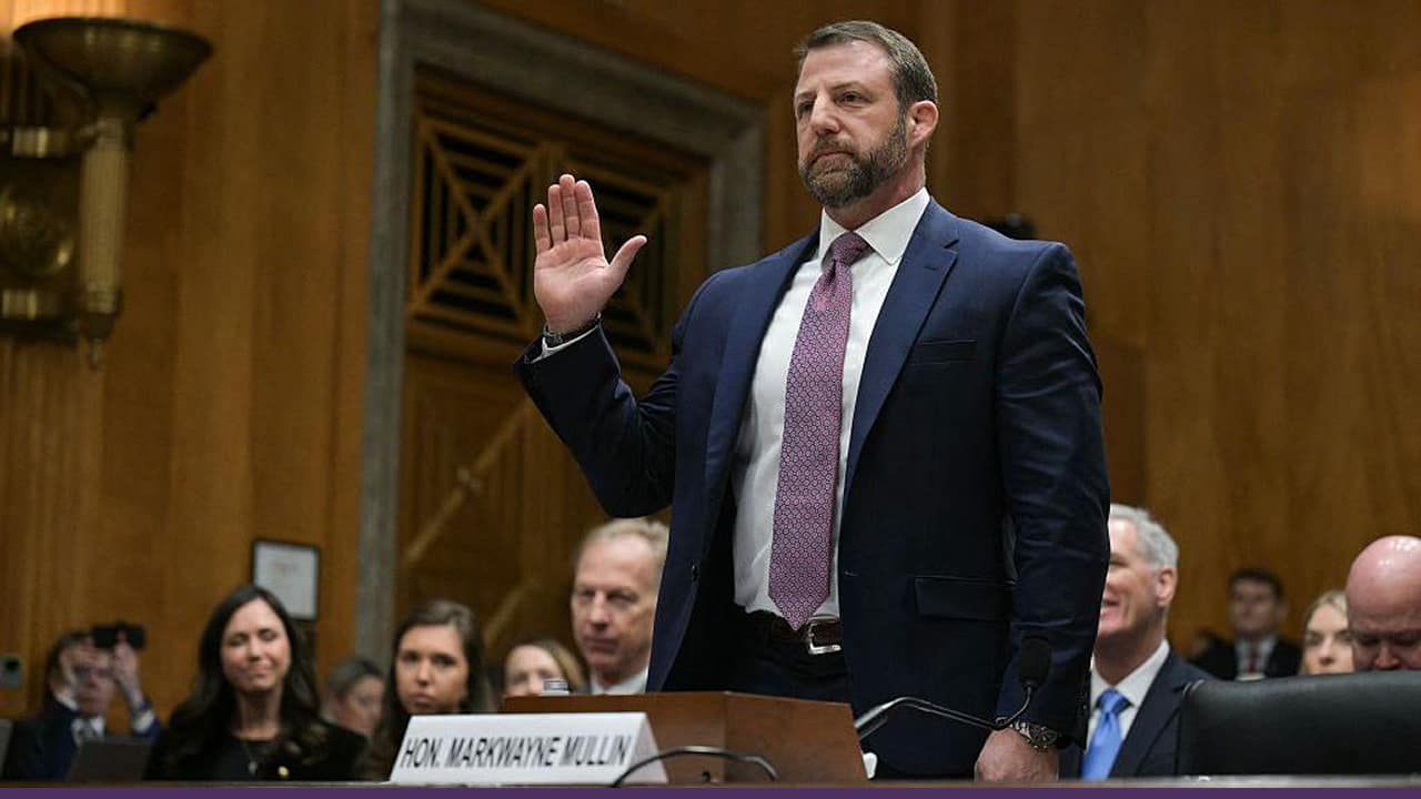 A man in a suit raises his right hand while being sworn in during a formal hearing, with seated officials and an audience visible in the background.