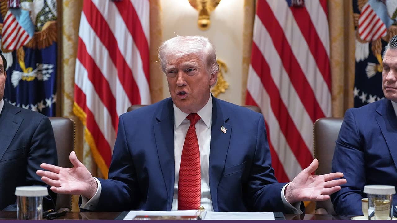 A man in a navy suit and red tie speaks while seated at a conference table, gesturing with both hands, with multiple U.S. flags displayed behind him and others seated on either side.