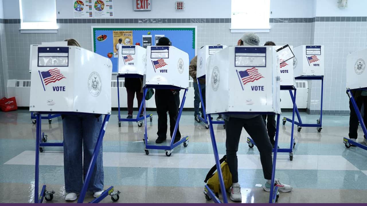 People casting their ballots at voting booths inside a polling station, each standing behind privacy screens marked with the U.S. flag and the word “Vote.”